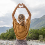 Eine blonde Frau ist von hinten zu sehen, formt mit den Händen über dem Kopf ein Herz und blickt auf einen Fluss, der von Wald und Bergen umgeben ist.